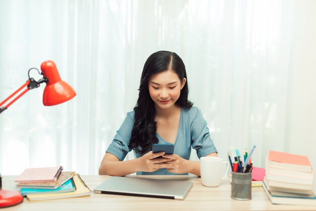 Belle jeune étudiante utilise un smartphone et prend des notes tout en étant assis au bureau