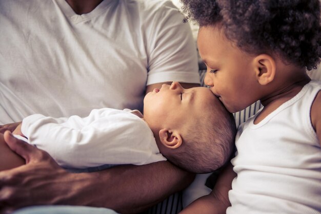 Belle jeune famille afro-américaine passant du temps ensemble. Le petit bébé dort dans les bras de papa pendant que sa soeur l'embrasse