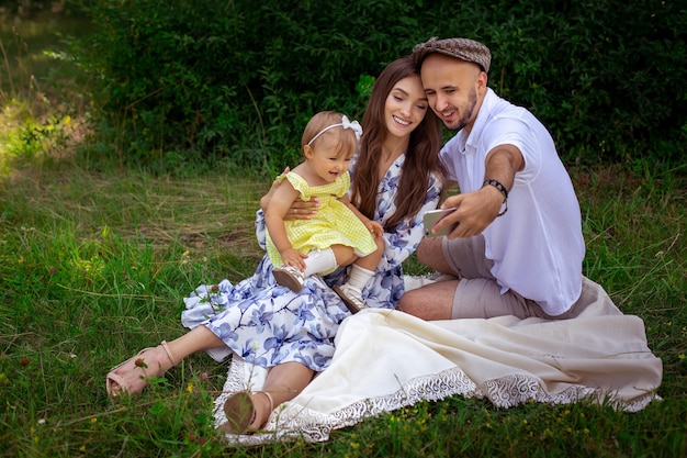 Belle jeune famille fait selfie sur pique-nique. Parents avec petite fille prenant une photo sur un téléphone portable dans le parc verdoyant