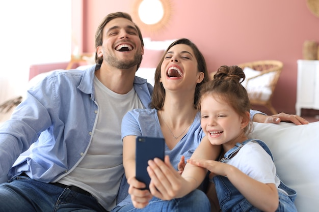 Belle jeune famille avec petit enfant prenant un selfie avec un smartphone à la maison sur le canapé.