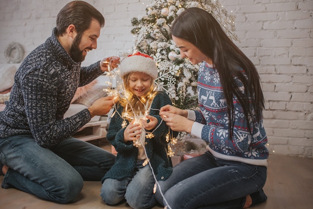 Belle jeune famille profitant de leurs vacances ensemble, décorant un sapin de Noël, arrangeant les lumières de Noël et s'amusant