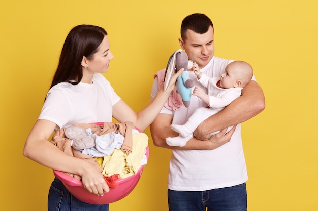 Belle jeune famille de trois personnes photographiée contre un mur jaune, maman faisant un landry et tenant un bassin plein de vêtements sales, père avec bébé dans les mains essayant de réconforter bébé, maman montre un jouet.
