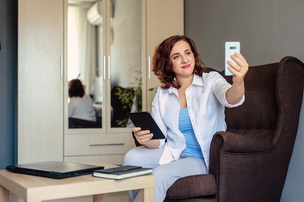 Belle jeune femme de 30 ans dans une chemise blanche fonctionne, assis sur une chaise dans le bureau à domicile et discute des problèmes avec le client à l'aide d'appels vidéo