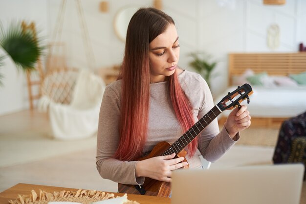 Une belle jeune femme accorde un ukulélé pour s'entraîner. Apprentissage à distance pour jouer d'un instrument de musique.