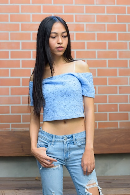 Belle jeune femme adolescente asiatique debout tout en pensant et regardant vers le bas contre le mur de briques