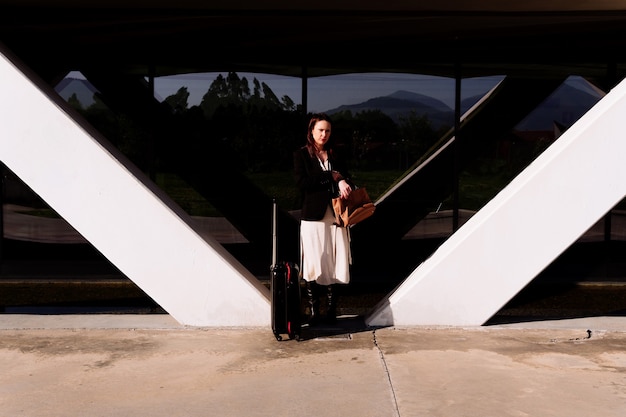 Belle jeune femme adulte en voyage d'affaires pensive dans la rue par quartier d'affaires ou parc industriel portant une valise. concept de voyage d'affaires