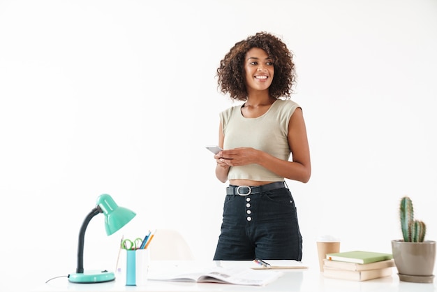 Belle jeune femme d'affaires africaine souriante portant des vêtements décontractés debout au bureau isolé sur un mur blanc, à l'aide d'un téléphone portable