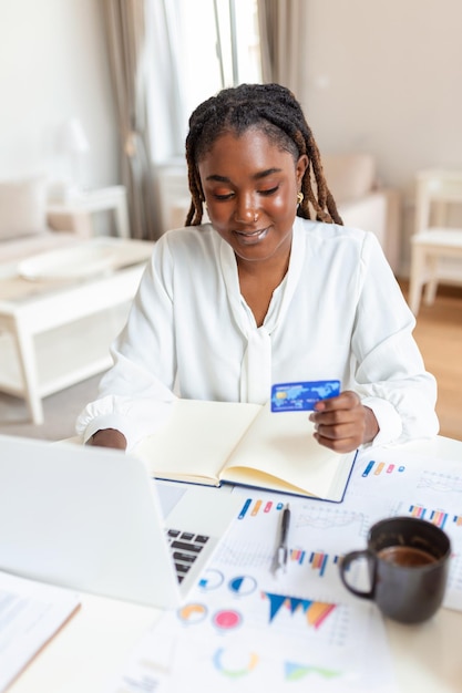 Belle jeune femme d'affaires africaine souriante tenant une carte de crédit et utilisant un ordinateur portable au bureau