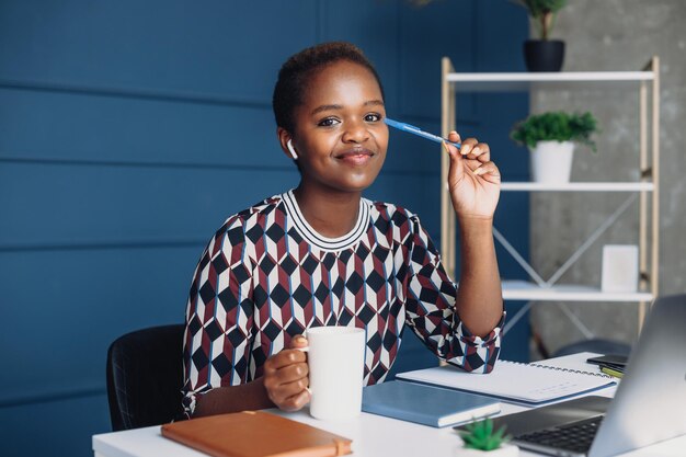 Photo belle jeune femme d'affaires africaine souriante tenant un stylo et regardant la caméra tout en travaillant avec un ordinateur portable au bureau concept d'entreprise d'emploi personne positive