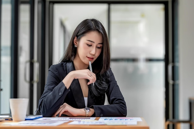 Belle jeune femme d'affaires asiatique tenant un stylo assis dans une chaise de bureau examinant le graphique des revenus du document placé sur la table.