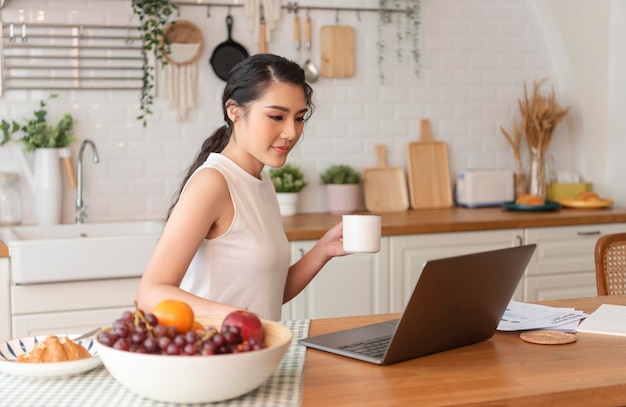 Belle jeune femme d'affaires asiatique travaillant dans la cuisine et buvant du café à la maison