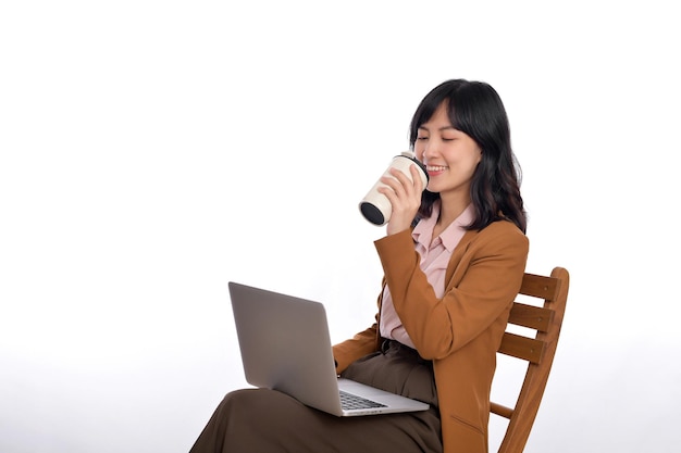 Belle jeune femme d'affaires asiatique utilisant un ordinateur portable assis sur une chaise blanche et buvant du café isoler sur fond blanc