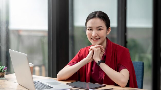 Belle jeune femme d'affaires asiatique vêtue d'une chemise rouge est heureuse de s'asseoir et de tablettes et d'un ordinateur portable au bureau. En regardant la caméra.