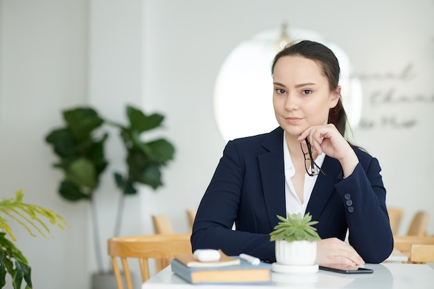 Belle jeune femme d'affaires confiante assise à la table du café, enlevant des lunettes et regardant la caméra