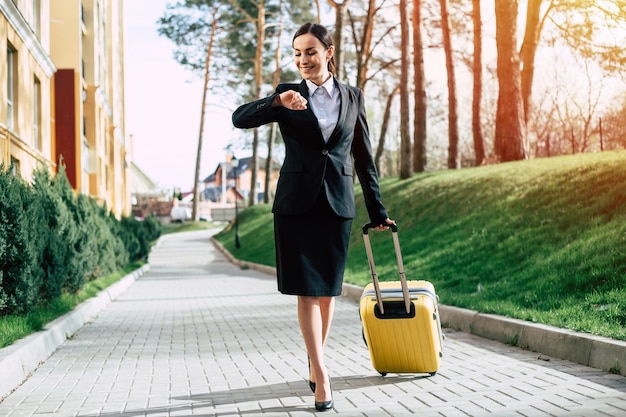 Belle jeune femme d'affaires en costume et chaussures marchant dans une rue de la ville avec une valise de voyage jaune