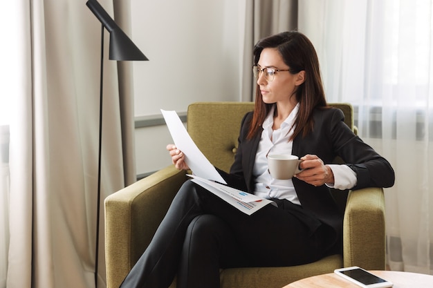 belle jeune femme d'affaires dans des vêtements de cérémonie à l'intérieur à la maison en buvant du café avec des documents.