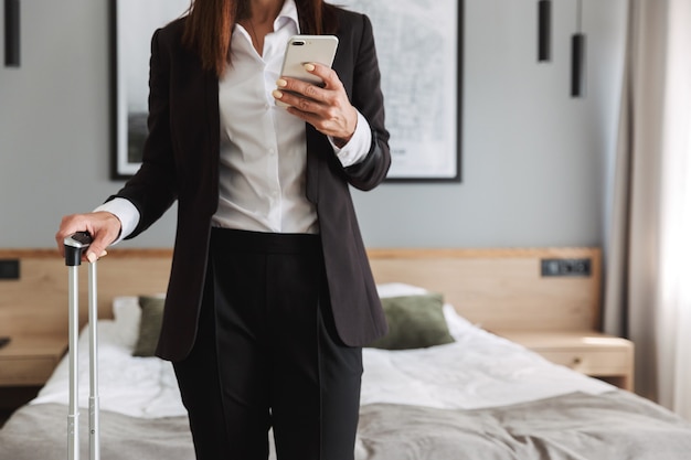 belle jeune femme d'affaires dans des vêtements de cérémonie à l'intérieur à la maison avec une valise à l'aide d'un téléphone portable.