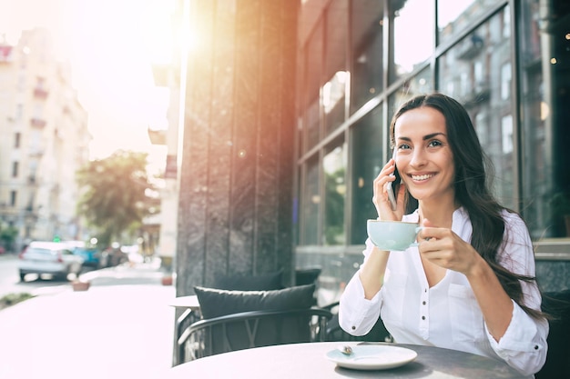 Belle jeune femme d'affaires mignonne souriante utilisant un smartphone et buvant du café dans un café d'été en plein air