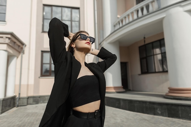 Belle jeune femme d'affaires à la mode avec des lunettes de soleil en vêtements de mode noirs avec un blazer noir et des promenades en haut et profiter de la ville