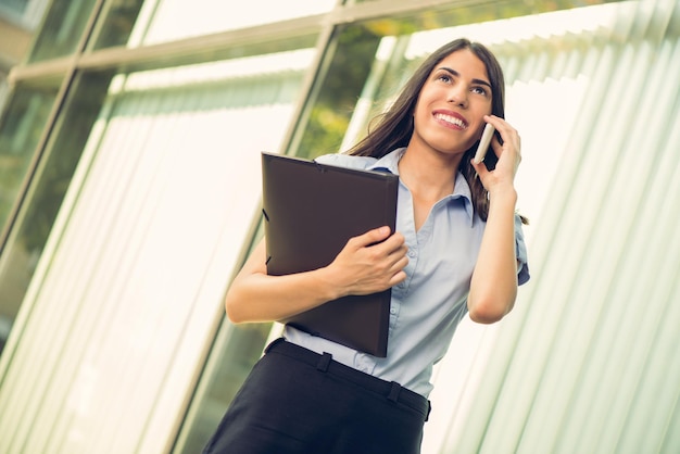 Belle jeune femme d'affaires avec un planificateur dans les mains, debout devant un immeuble de bureaux et parlant au téléphone mobile