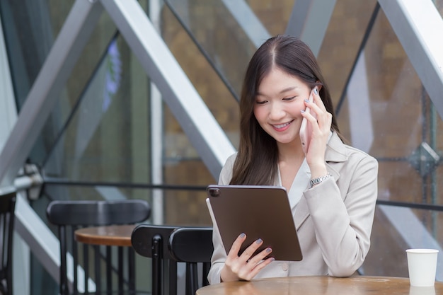 Belle jeune femme d'affaires professionnelle asiatique dans un costume de couleur crème tient un smartphone pour parler, discuter et enregistrer des informations joyeusement sur une tablette dans un café.