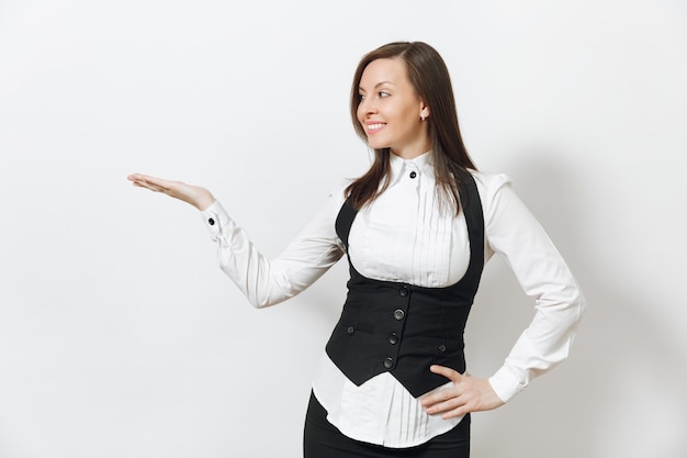 Belle jeune femme d'affaires souriante aux cheveux bruns en costume noir, chemise blanche, pointant la main droite de côté isolé sur mur blanc