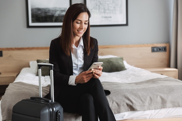 belle jeune femme d'affaires souriante heureuse dans des vêtements de cérémonie à l'intérieur à la maison avec une valise à l'aide d'un téléphone portable.