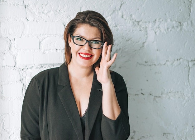 Belle jeune femme d'affaires souriante à lunettes et veste noire contre un mur blanc