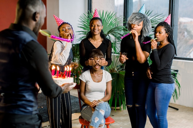 Belle Jeune Femme Africaine Aux Cheveux Noirs, Assise Sur La Chaise Avec Ses Amis Autour D'elle, Célébrant Et Soufflant Ses Bougies D'anniversaire. Un Africain Porte Un Gâteau D'anniversaire Avec Des Bougies