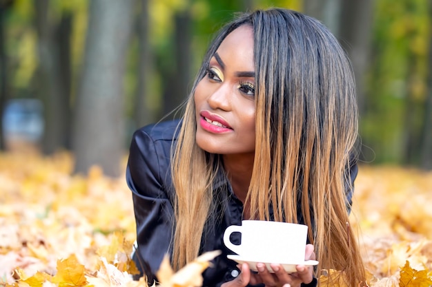Belle jeune femme africaine de boire du café dans une tasse blanche sur les feuilles d'automne dans le parc
