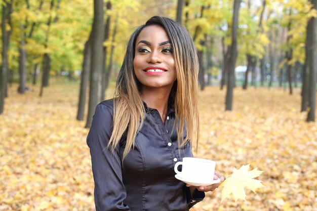 Belle jeune femme africaine de boire du café dans une tasse blanche sur le fond des feuilles d'automne dans le parc