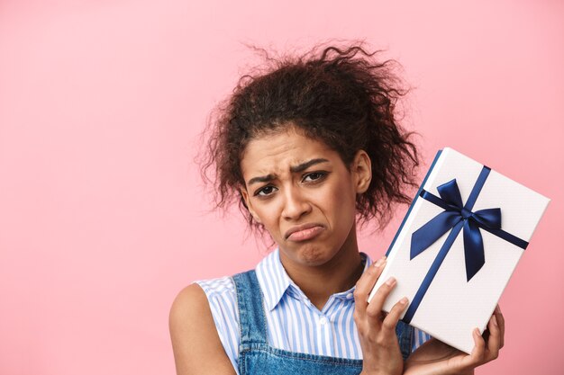 Belle jeune femme africaine déçue tenant une boîte-cadeau sur rose