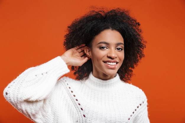 Belle jeune femme africaine joyeuse portant un pull debout isolé sur un mur rouge, essayant d'entendre des rumeurs