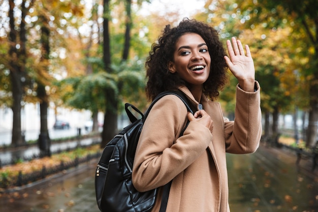 Belle jeune femme africaine portant un manteau marchant à l'extérieur dans le parc, portant un sac à dos