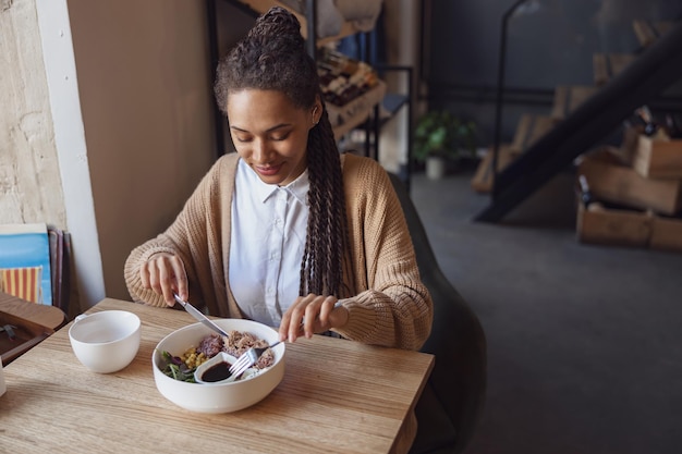 Belle jeune femme afro-américaine est en train de déjeuner dans un restaurant en dégustant un plat sain servi dans un bol blanc