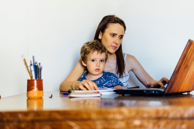 Belle jeune femme d'âge moyen à la maison assise à une table avec un ordinateur avec son fils de trois ans pris.concept télétravail.maternité. conciliation