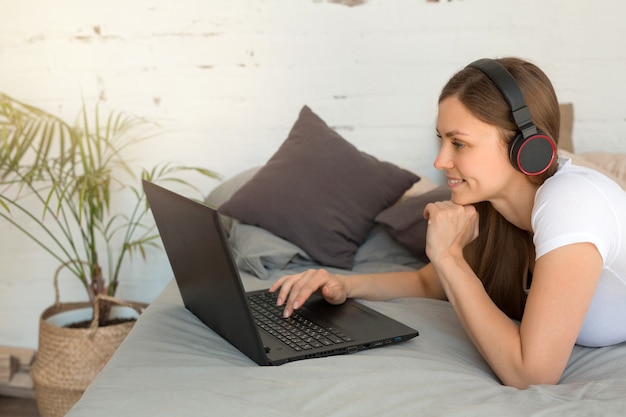 Belle jeune femme allongée sur le lit avec un ordinateur portable à la maison dans les écouteurs