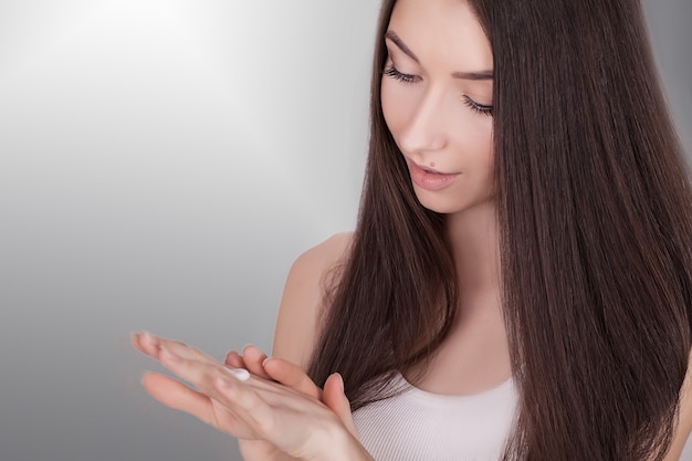 Belle jeune femme appliquant la crème pour le visage sur son pommette dans un soin de la peau ou des cosmétiques sur fond gris avec espace de copie