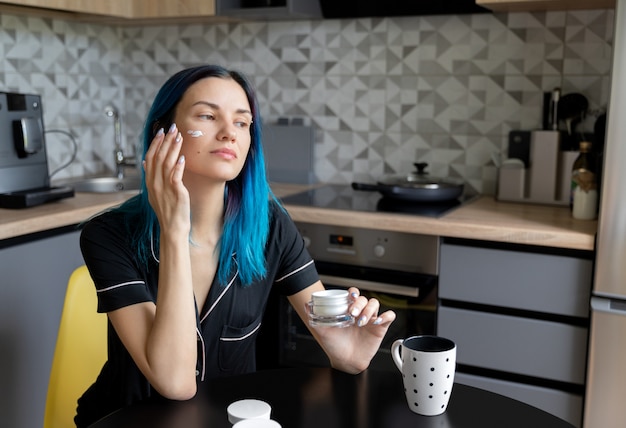 Belle jeune femme appliquant de la crème sur son visage à la cuisine moderne