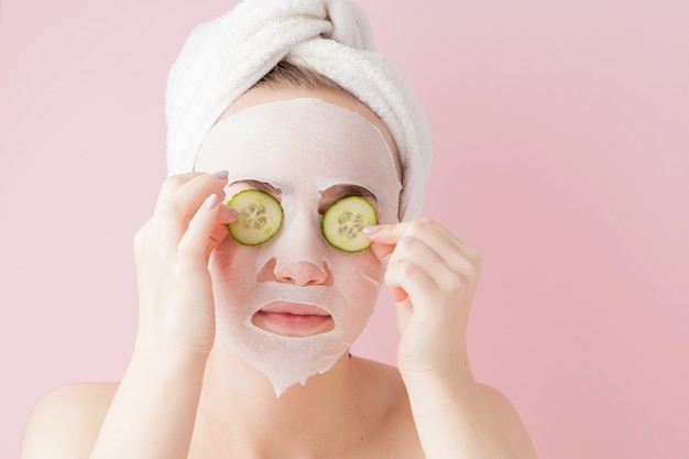 Belle Jeune Femme Applique Un Masque De Tissu Cosmétique Sur Un Visage Avec Du Concombre Sur Fond Rose. Soins De Santé Et De Beauté Et Concept Technologique