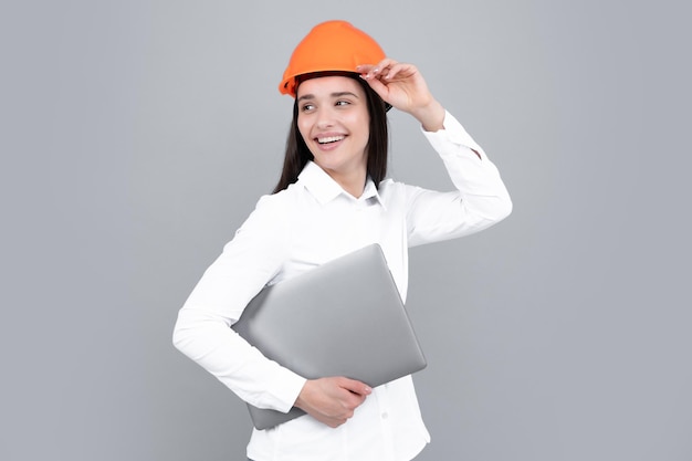 Belle jeune femme architecte en casque de casque tenir un ordinateur portable sur fond gris Constructeur féminin en casque pour enseigne publicitaire