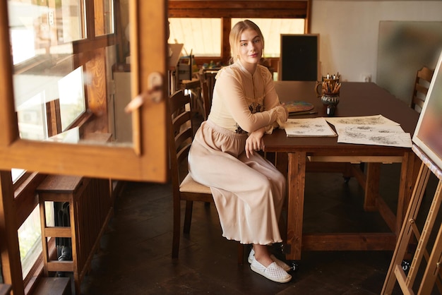 Une belle jeune femme artiste ou poète créative est assise à une table en bois dans un studio d'art et écrit une image ou une lettre à l'encre. Photo élégante romantique dans des tons beiges chauds