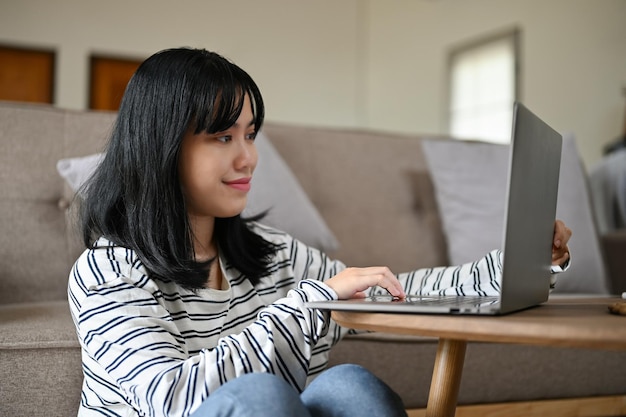 Belle jeune femme asiatique à l'aide de son ordinateur portable dans son salon à la maison