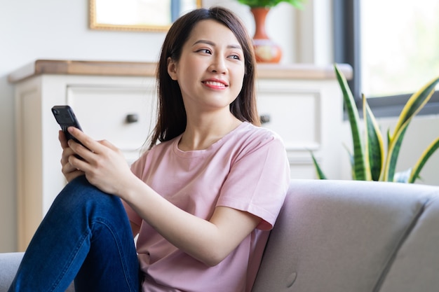 Belle jeune femme asiatique assise sur un canapé à l'aide de son téléphone et regardant par la fenêtre