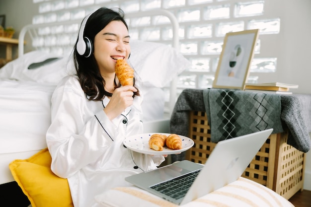 Belle jeune femme asiatique assise à côté du lit à l'aide d'un ordinateur portable et petit-déjeuner le matin à la maison