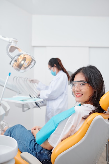 Belle jeune femme asiatique assise dans un fauteuil dentaire et souriant à la caméra, dentiste choisissant des pièces à main en arrière-plan