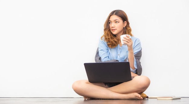 Belle jeune femme asiatique assise sur le sol tenant une tasse de café tout en prenant une pause de travailler sur son ordinateur à la maison