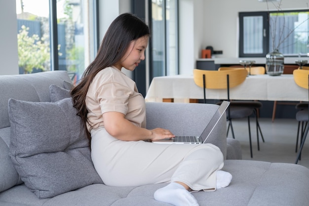 belle jeune femme asiatique dans des vêtements confortables et décontractés à l'aide d'un ordinateur portable assis sur un canapé dans un appartement moderne