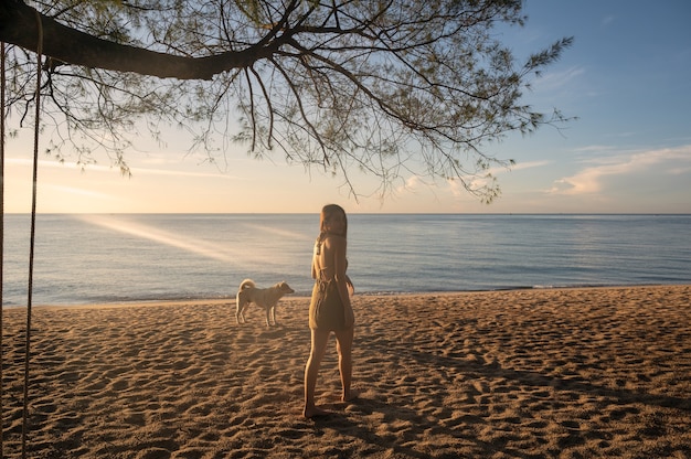 Belle jeune femme asiatique debout avec un chien errant et du soleil sur la plage le matin