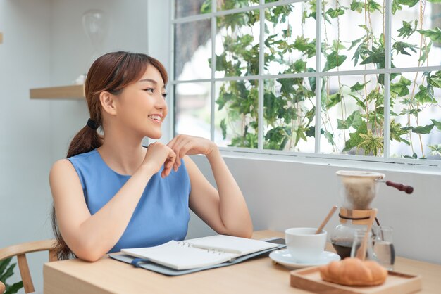 Belle jeune femme asiatique écrivant pour faire la liste tout en étant assis à la fenêtre